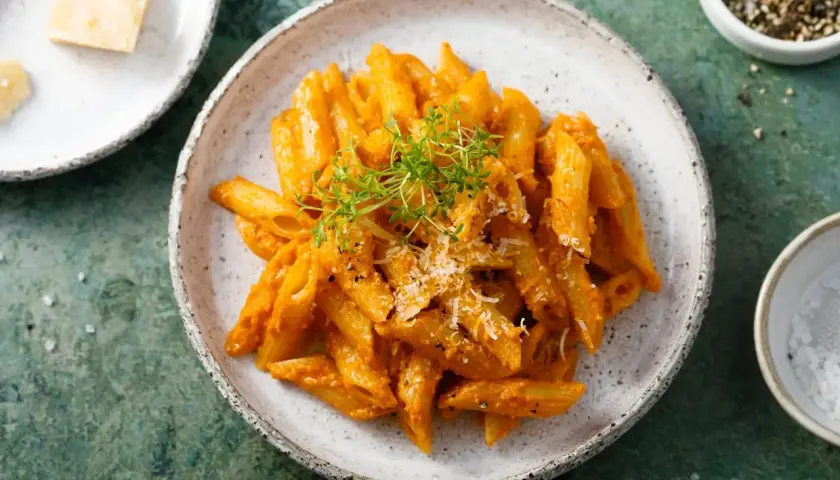 Penne z aromatycznym, czerwonym pesto, posypane parmezanem i świeżymi ziołami. Obok miseczka z solą i pieprzem.