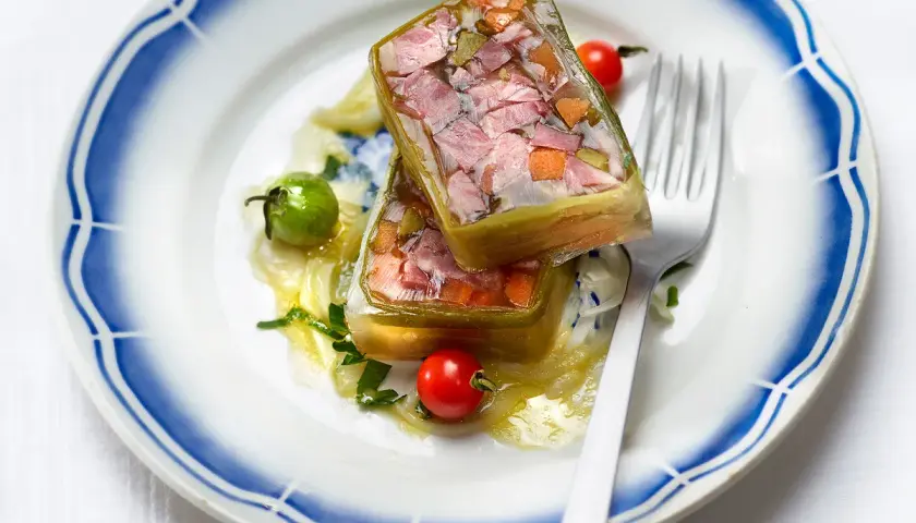 Deux portions de fromage de tête translucide, garnies de légumes et de viande, sur une assiette ornée de bleu.