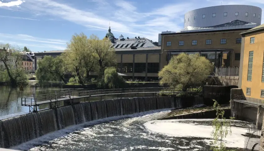 Vattenfall porlar förbi Louis de Geer Konsert & Kongress, en modern byggnad med en rundad takkonstruktion, omgiven av grönska och gamla industribyggnader.
