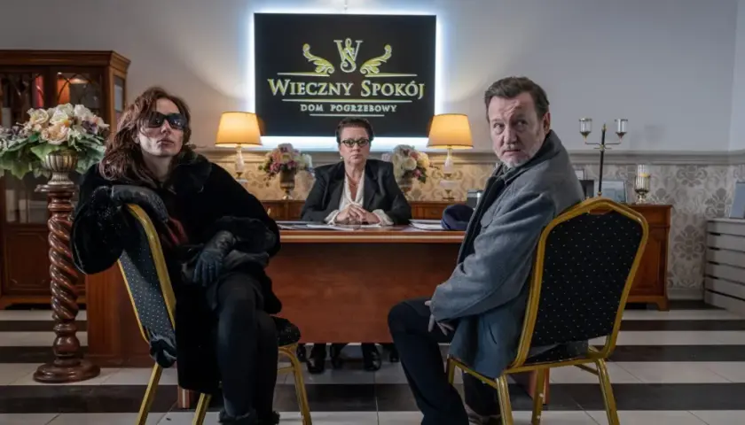 Trzy osoby siedzą w eleganckim biurze domu pogrzebowego. W tle logo "Wieczny Spokój". Scena z serialu "Minuta ciszy sezon 2".