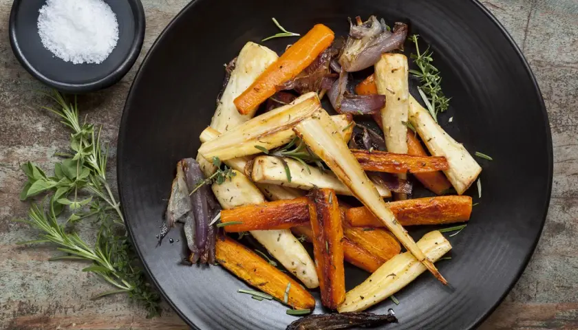 Légumes rôtis, carottes et panais, parsemés d'herbes. Un navet doré, tendre et savoureux, se mêle aux autres légumes sur l'assiette noire.