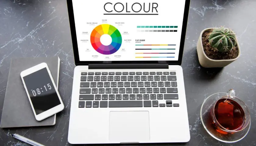 Na ekranie laptopa widać koło barw i palety kolorów, prezentujące różnice między cmyk vs rgb. Obok telefon, notes i filiżanka herbaty.