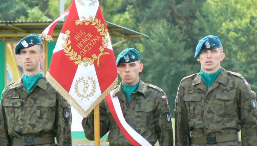 Trzech żołnierzy rezerwy w mundurach i beretach stoi obok sztandaru.
