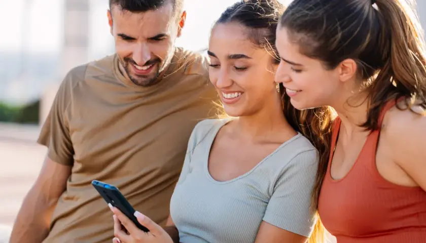 Trzech przyjaciół ogląda coś na telefonie. Uśmiechnięta dziewczyna, która trzyma telefon, może ukrywać zakochanie, gdy patrzy na ekran.