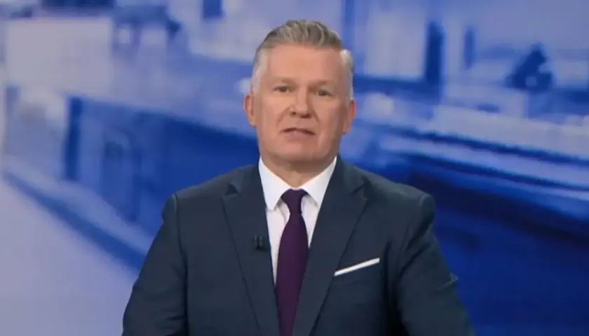 Jeden z prezenterów Fakty TVN w granatowym garniturze i bordowym krawacie, na tle niebieskiej grafiki.