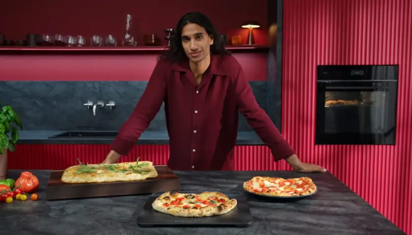 Mann in bordeauxrotem Hemd präsentiert frisch gebackene Pizzen. Im Hintergrund ein Ofen mit Pizzafunktion, der eine weitere Pizza bäckt.