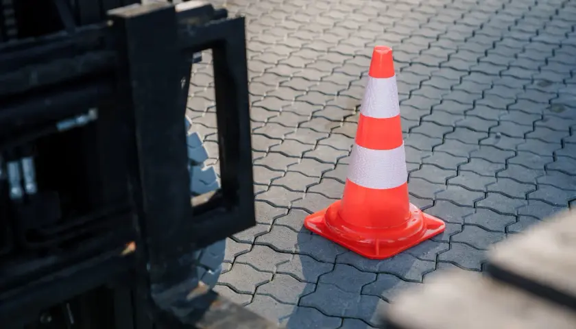 Ein orange-weißer Verkehrskegel steht auf einem gepflasterten Boden neben einem Gabelstapler. Man fragt sich, ob ein Staplerschein ablaufen kann.