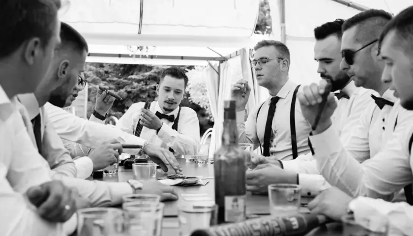 Panowie w eleganckich koszulach i szelkach świętują kawalerski pomysły przy cygarach i drinkach.
