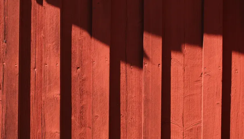 Röd träfasad med skuggor som faller som vertikala streck. Träet är målat tunt, vilket ger en rustik känsla.