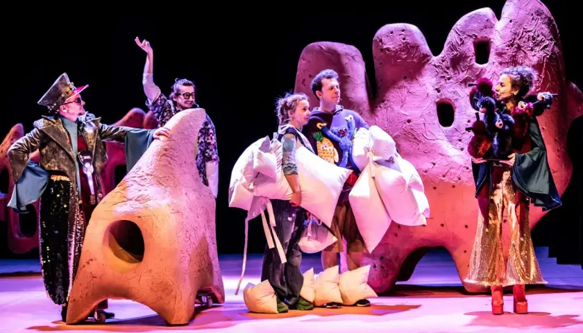 Aktorzy w barwnych strojach, niosący poduszki, na tle scenografii przypominającej gigantyczne, różowe formacje. To fragment spektaklu z repertuaru teatru lalki.