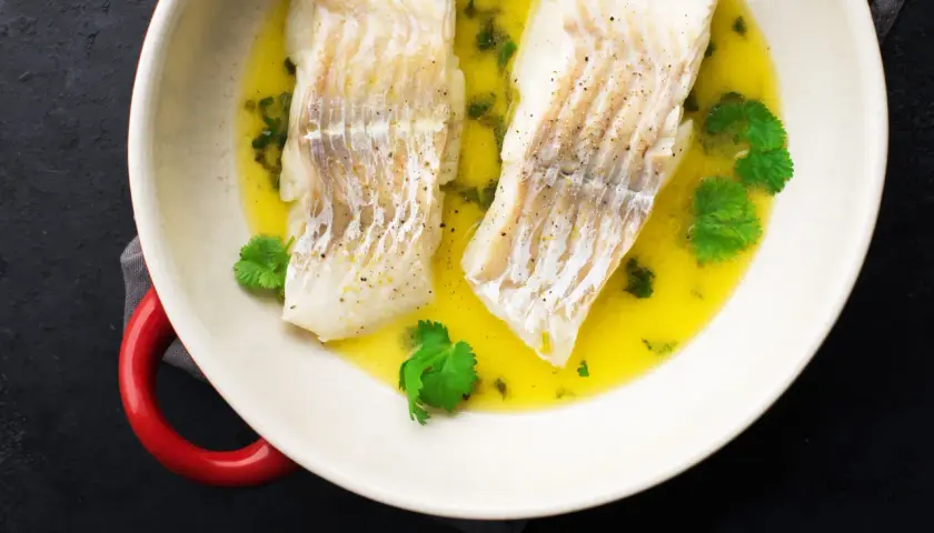 Dwa filety halibut pieczony ze skórą w maśle z ziołami i kolendrą, podane w białej misie z czerwonymi uchwytami.