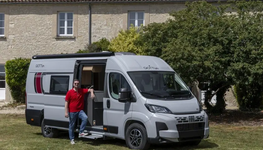 Un homme pose devant un **fourgon ou camping car** gris, porte ouverte, devant une maison en pierre et des arbres.