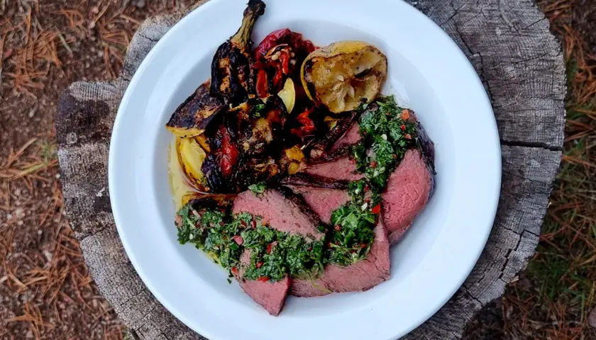 Skivad picanha med perfekt picanha temp, toppad med chimichurri och grillade grönsaker. Serveras på en vit tallrik på en trädstam.