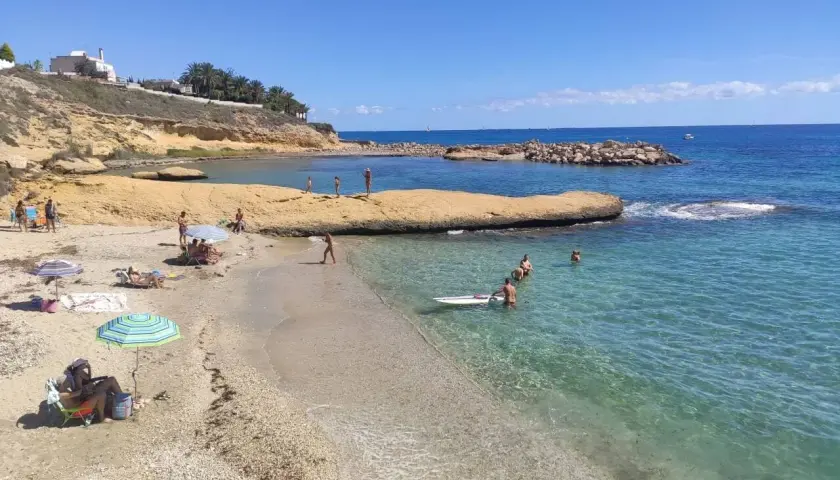 Gente disfrutando del sol y el mar en la cala Almadraba, Campello. Aguas cristalinas y rocas emergiendo del azul.