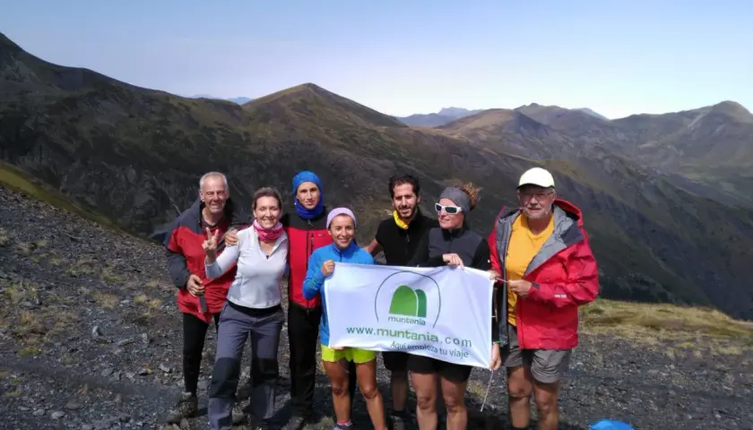 Grupo de senderistas celebra en la cima de una montaña, con el valle de **Aran** de fondo. Sostienen una pancarta de Muntania.com.