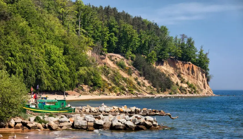 Zielona łódź rybacka przy kamienistym brzegu, na tle zielonego lasu i piaszczystej plaży. W oddali widać wysokie, strome urwisko porośnięte drzewami.