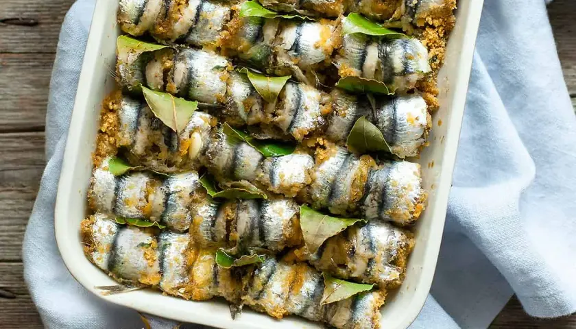 Sarde a beccafico pronte per essere infornate, disposte in una teglia con foglie di alloro e panatura dorata.