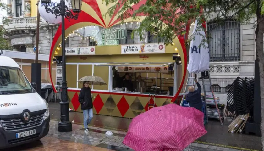 Preparativos para las fiestas de San Mateo en Oviedo. Un puesto de comida y bebida con decoración festiva, gente con paraguas y un furgón blanco.