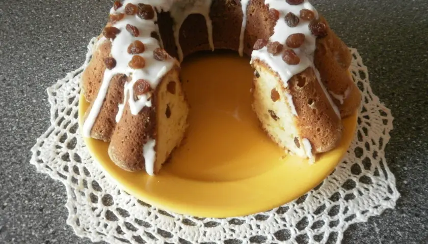 Pyszna babka drożdżowa Ani gotuje, polana lukrem i posypana rodzynkami, na żółtym talerzu.