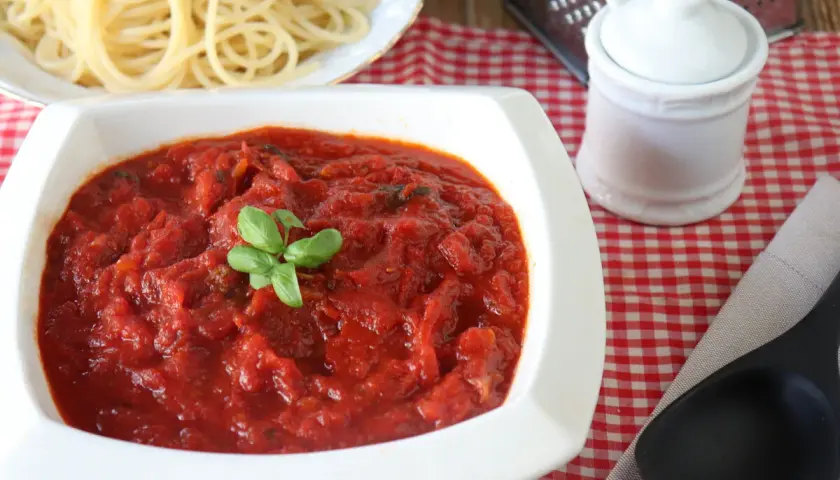 Un tazón blanco lleno de salsa marinara casera, decorada con albahaca fresca, junto a espaguetis cocidos.