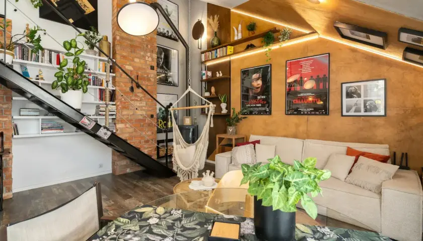 Wnętrze w stylu loftowym z ceglaną ścianą, metalowymi schodami, wiszącym fotelem i wygodną kanapą.