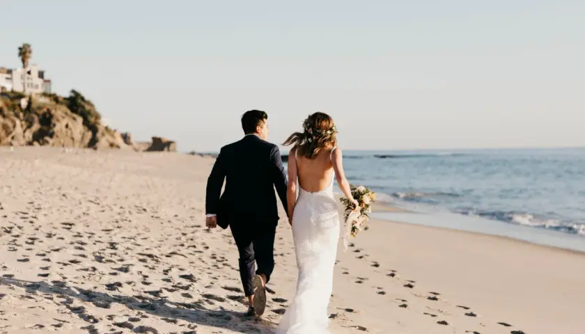 Para nowożeńców spaceruje po plaży. Panna młoda w pięknej sukni ślubnej z bukietem kwiatów, pan młody w granatowym garniturze. Słońce zachodzi nad oceanem.