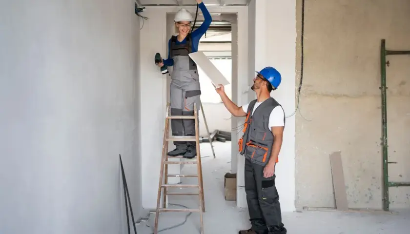 Dwóch budowlańców w kaskach pracuje przy suficie. Kobieta na drabinie trzyma wiertarkę, mężczyzna podaje jej płytę gipsowo-kartonową. W tle widać instalacje i materiały budowlane, jakby przygotowywali się do montażu **fizeliny**.