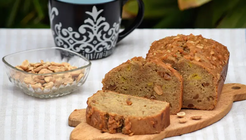 Un delicioso bizcocho de plátano, cubierto de frutos secos, acompañado de cacahuetes y una taza.