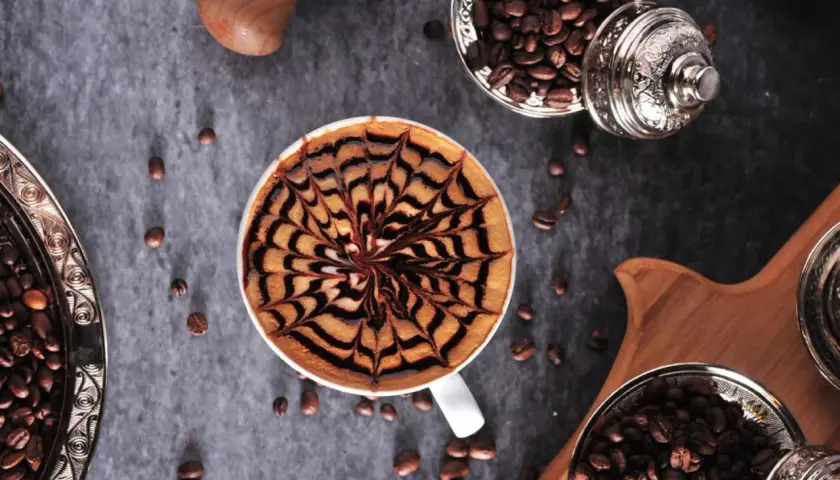 Kawa mocha z artystycznym wzorem na piance. Obok ziarna kawy w ozdobnych naczyniach.