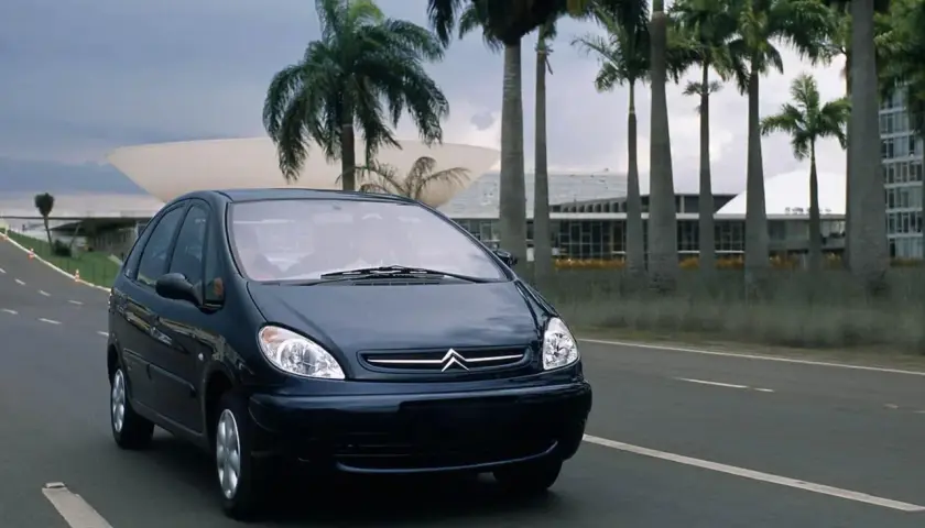 Ciemnoniebieski Citroen Xsara Picasso jedzie drogą wśród palm, w tle nowoczesna architektura.