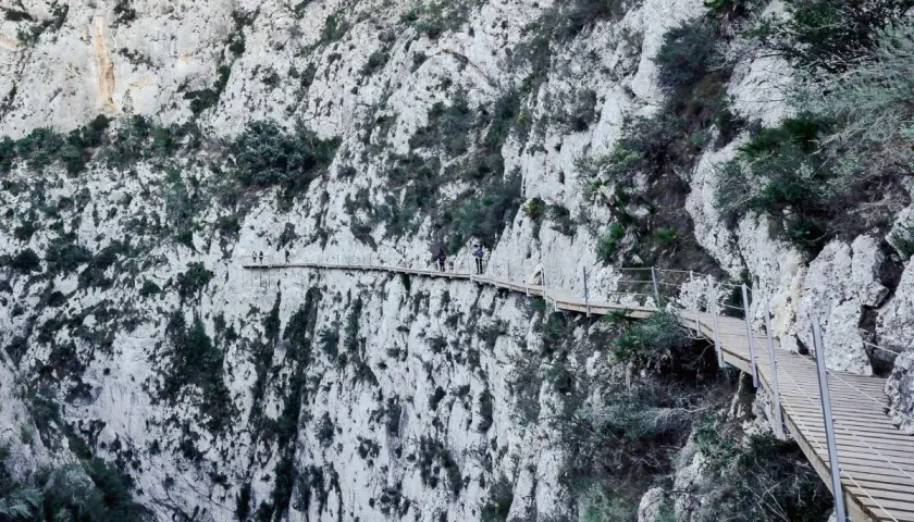 Personas caminan por la **ruta de las pasarelas**, un sendero de madera colgado en un acantilado rocoso con vegetación.