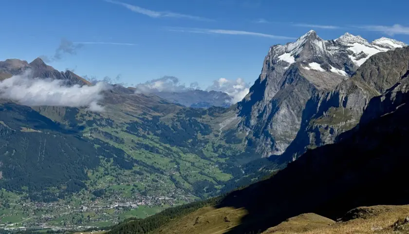 Panoramic view of a valley village nestled among green hills, with dramatic, snow-capped peaks of the Swiss Alps in the background. This scene exemplifies the best hikes in Swiss Alps.