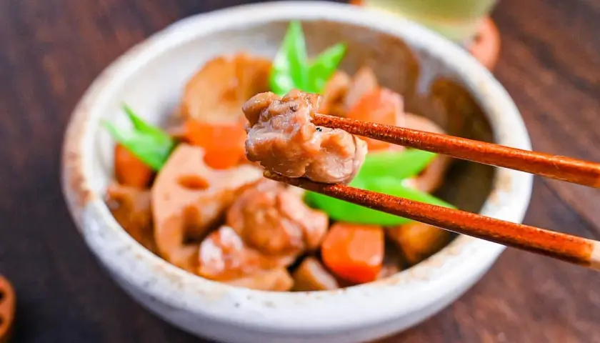 A close-up of a bowl of chikuzenni recipe, with chopsticks lifting a piece of chicken. The dish contains chicken, lotus root, carrots, and green beans.