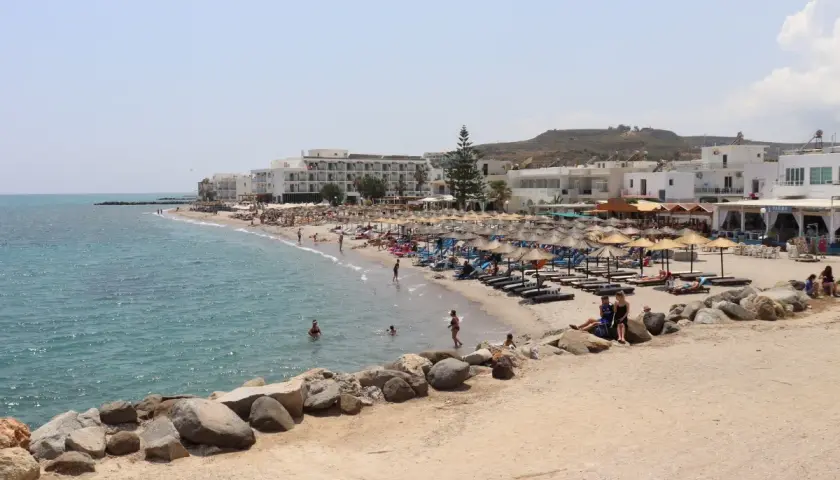 Słoneczna plaża na wyspie Kos z białymi hotelami, parasolami i turystami kąpiącymi się w morzu.