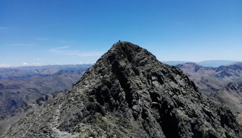 Cima rocosa de montaña con excursionistas y vista panorámica de picos bajo un cielo azul.