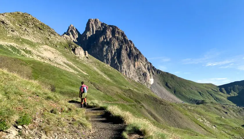 Senderista se dirige hacia el imponente Midi d'Ossau, rodeado de verdes laderas y un cielo azul despejado.