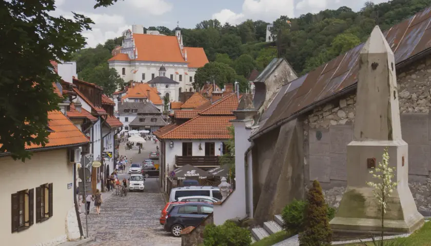 Ulica w Kazimierzu Dolnym z widokiem na kościół i wzgórza. Widać kamienne domy, brukowaną drogę i turystów. Kazimierz Dolny atrakcje zachwycają.
