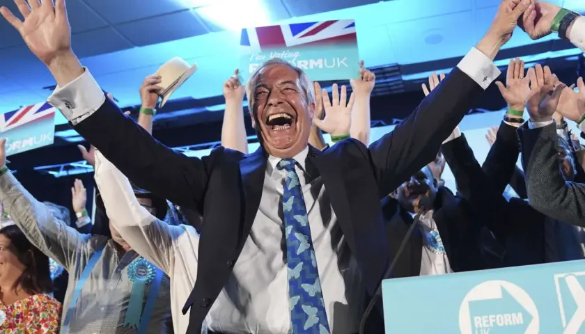 Jubelnde Menge bei einer Wahl in England. Ein Mann mit erhobenen Armen und einem breiten Lachen im Vordergrund, umgeben von Union-Jack-Flaggen und "Reform UK"-Schildern.