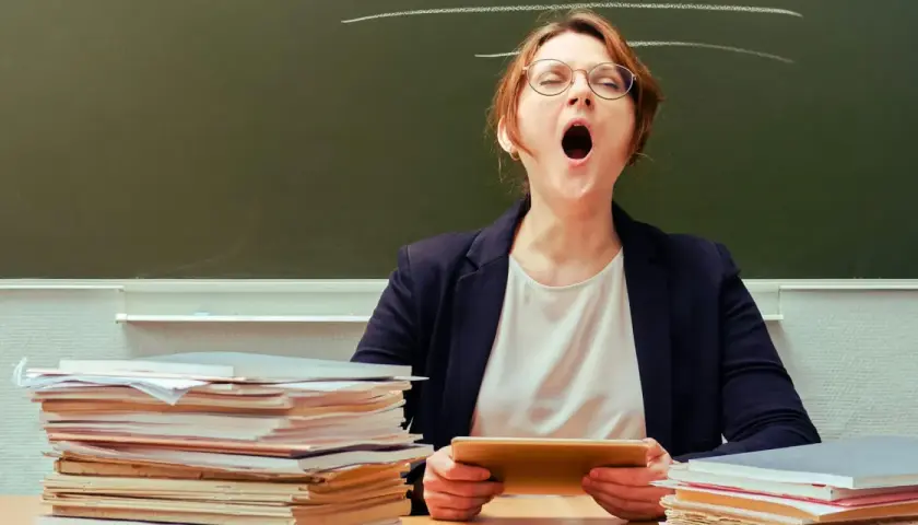 Lehrerin mit Brille gähnt erschöpft hinter einem Stapel Bücher. Ein Klischee, das viele kennen.
