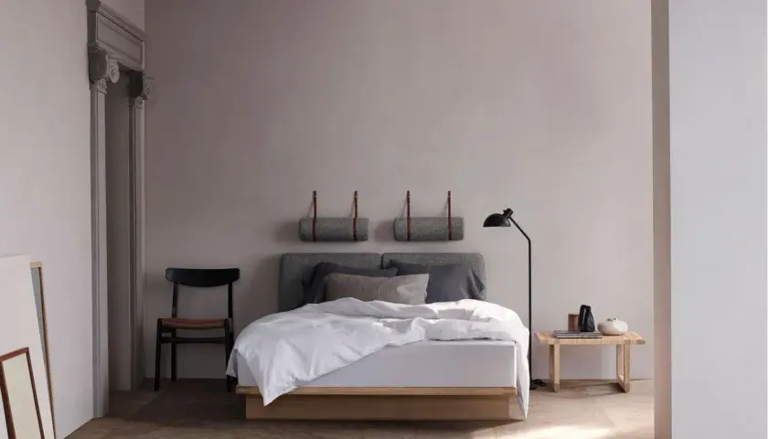Ein minimalistisches schlafzimmer skandinavisch mit einem Holzbett, grauen Kissen und einer Stehlampe. Zwei gerollte Decken hängen an der Wand.