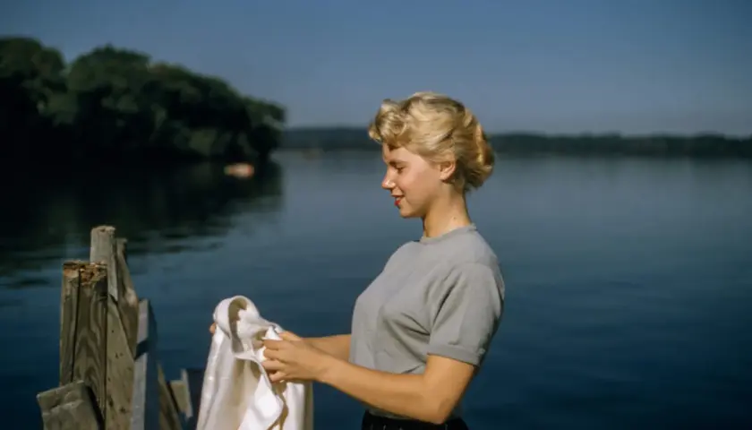 Joven con peinados años 50, con un suéter gris y falda a cuadros, sostiene una chaqueta blanca junto a un lago.