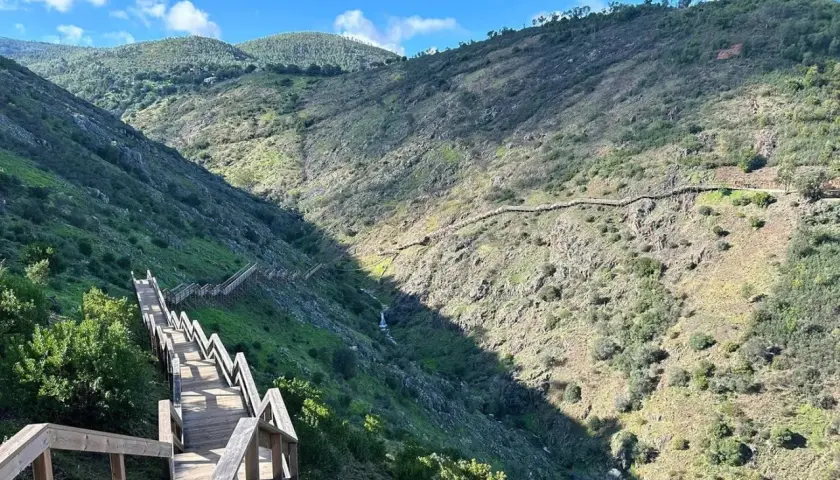 Senderismo Algarve: escaleras de madera descienden por un barranco verde y rocoso, con un sendero serpenteante en la ladera opuesta.