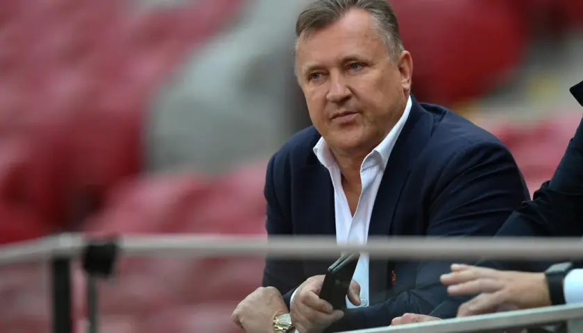 Prezes PZPN w garniturze, z telefonem w dłoni, siedzi na trybunach stadionu.