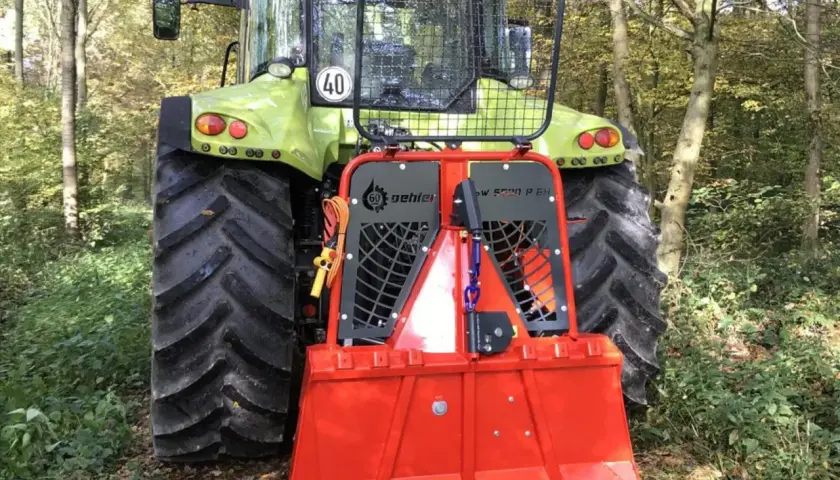 Ein grüner Traktor mit einer roten Forstseilwinde im Wald. Die Seilwinde ist bereit für den Einsatz, um Holz zu ziehen.