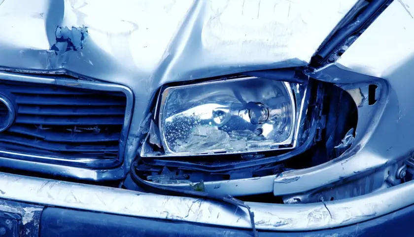 Il cofano e il faro di un'auto sono gravemente danneggiati, con lamiera accartocciata e vetro rotto. Sembra il risultato di **atti vandalici**.