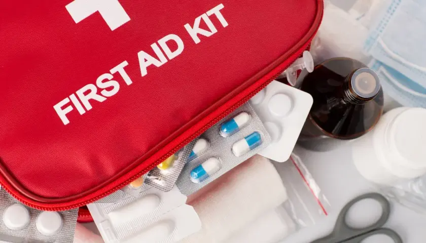 Czerwona apteczka z białym krzyżem i napisem "FIRST AID KIT", zawierająca leki, bandaże i nożyczki.