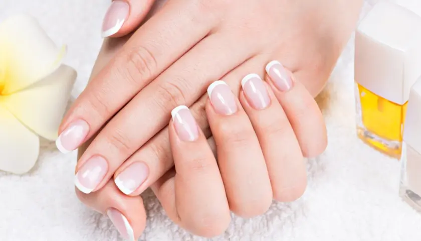 Elegancki manicure french z diamencikami, dłonie zadbane i wypielęgnowane, obok kwiat i lakiery.