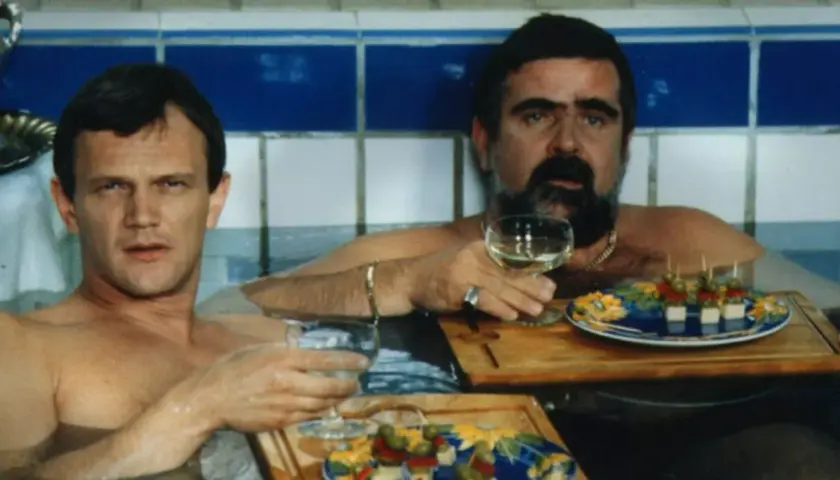 Dwóch mężczyzn w wannie, jeden z wąsami, pije wino i je przekąski. Scena z filmu "Kiler", rok produkcji 1997.
