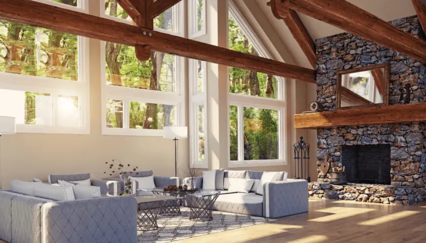 Vaste salon avec cheminée en pierre et un impressionnant plafond cathédrale bois. De grandes fenêtres donnent sur une forêt luxuriante.