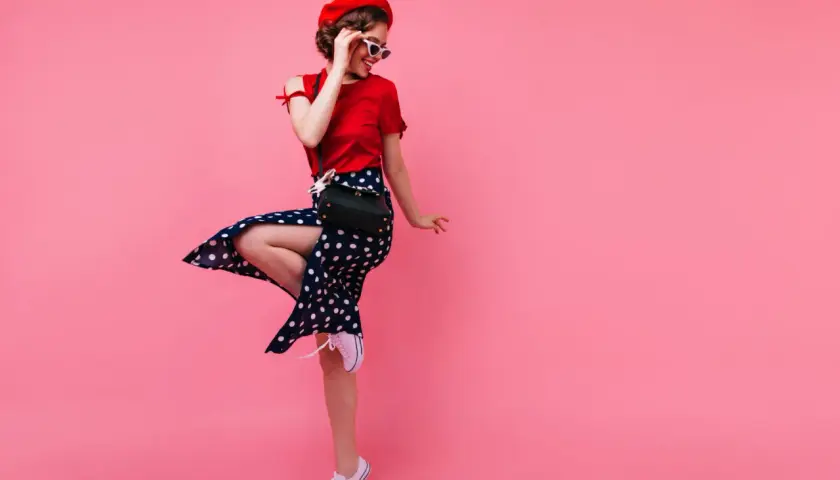 Dziewczyna w stylu pin-up w czerwonej bluzce i spódnicy w grochy tańczy na różowym tle.